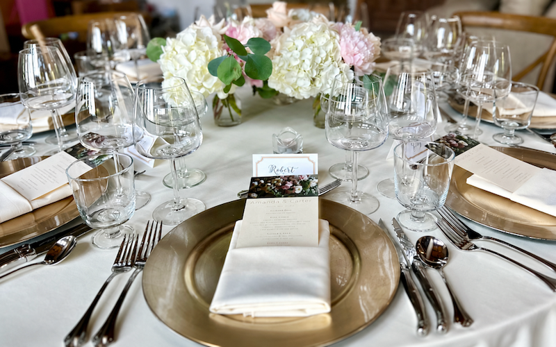 Dining table set with gold chargers, white linen napkins, wine glasses, and a centerpiece of white hydrangeas and pale pink flowers.